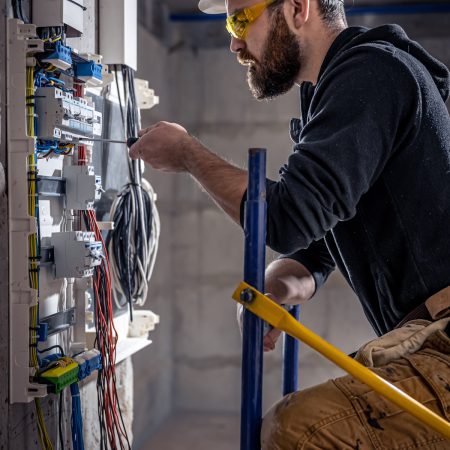 A male electrician works in a switchboard with an electrical connecting cable, connects the equipment with tools.